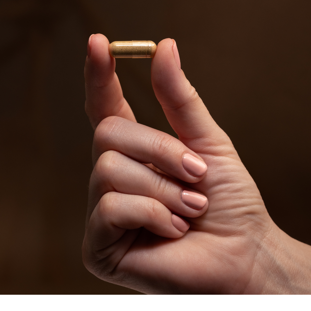 Close-up of a hand holding a single Grass-Fed Beef Liver supplement capsule, highlighting its natural brown color and compact size, against a warm, neutral background.